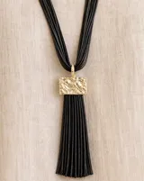 Collar largo con tira de cuero negro y dije metálico dorado rectangular del que cuelga un gran fleco de hilos negros.