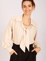Blusa de manga larga color beige claro, con cuello camisero y lazo frontal ajustable. Presenta puños fruncidos con elástico.
