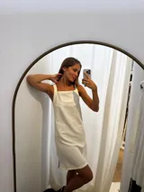 Vestido corto de lino en color beige, con escote cuadrado y breteles anchos. Presenta pespuntes decorativos y tajos laterales para mayor comodidad y movimiento.