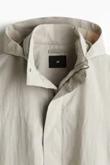 Campera parka de nylon color beige claro, con cuello elevado que oculta una capucha desplegable. Presenta cierre frontal oculto por solapa con botones a presión, bolsillos con cierre y bolsillos de solapa con botones a presión en la parte delantera, y un bolsillo interior.