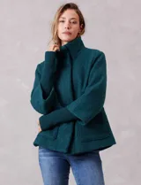 Tapado de tejido bouclé color marrón, con silueta evasé y largo a la cadera. Presenta cuello alto envolvente, cierre frontal oculto con botones a presión y bolsillos frontales de gran tamaño. Sus mangas son 3/4 con corte amplio y puños de tejido rib al tono que se extienden para cubrir las muñecas.