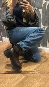 Bota tipo Chelsea de cuero marrón con suela track negra de goma. Presenta paneles elásticos laterales y una costura reforzada en la puntera. Incluye una correa de cuero desmontable con hebilla metálica.