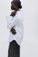 Camisa blanca de popelina de algodón, corte oversized con cuello clásico, hombros caídos y mangas largas con puños anchos abotonados. Presenta cierre frontal de botones y bajo redondeado.
