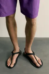 Sandalia multitiras de hombre, con suela plana tostada y tiras de tejido negro con costuras amarillas, ajuste con hebilla plástica y separación para el dedo.
