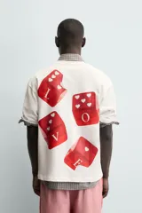 Camiseta blanca de corte relajado (relaxed fit) con cuello redondo y manga corta. Presenta un estampado grande en la espalda que simula dados rojos con letras y corazones.