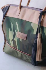 Bolso de fin de semana estilo tote de lona con estampado camuflado, detalles en cuero marrón y gamuza azul, manijas largas y borla decorativa colgante.