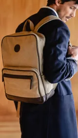 Mochila beige de tela canvas combinada con detalles en símil cuero marrón. Tiene apertura superior con cierre, bolsillo frontal con cierre, bolsillos laterales, bolsillos internos, asa corta de mano y correas acolchonadas regulables para la espalda.