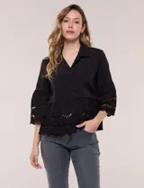 Camisa negra de poliéster con cuello camisero, escote en V y cierre frontal de botones. Cuenta con mangas anchas de corte acampanado y detalles de bordado suizo calado en las mangas y en el ruedo inferior.