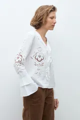 Blusa blanca de manga larga con diseño calado y bordado floral. Presenta un escote en V con detalle de volados frontales y mangas acampanadas con terminación en volado.
