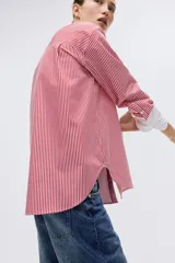 Camisa oversized de popelina de algodón con diseño a rayas verticales rojas y blancas. Presenta cuello clásico y puños anchos en color blanco sólido, cierre frontal con botones y bajo redondeado con aberturas laterales.