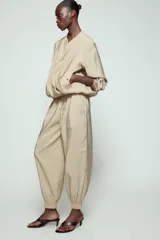 Pantalón satinado de tiro alto color beige, con cintura elástica ajustable con cordones, bolsillos delanteros y puños elásticos en los tobillos.
