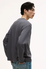 Remera de manga larga color gris oscuro, confeccionada en punto de algodón de peso medio. Presenta cuello redondo acanalado, hombros caídos, puños acanalados y bajo recto. Tiene un corte holgado.