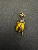 Dije en forma de escarabajo con acabado dorado envejecido, decorado con incrustaciones de cristales brillantes en la cabeza y el centro del cuerpo.