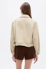 Chaqueta corta de algodón color beige, con cuello camisero, bolsillos laterales y cierre frontal con botones oscuros.