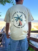 Remera oversize de color beige con estampa en la espalda de una palmera y una tabla de surf, acompañada de la inscripción "Surfer's Paradise Laguna Beach".