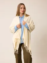 Pashmina de textura suave y color crema, con diseño liso y terminaciones en flecos en los extremos. Incluye una etiqueta decorativa en una de las esquinas.