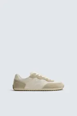 Zapatilla deportiva estilo barefoot de color beige, confeccionada en una combinación de materiales textiles y detalles en gamuza. Presenta puntera redondeada y ancha, plantilla OrthoLite extraíble y suela de goma fina, flexible y ligera en contraste.