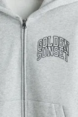 Campera con capucha de felpa gris, con cierre frontal completo, bolsillos tipo canguro y estampado tipográfico en el pecho. Presenta hombros caídos y puños acanalados.