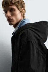 Cazadora negra de algodón con efecto lavado, corte relajado y capucha ajustable. Presenta cierre frontal de cremallera, bolsillos frontales tipo canguro y elásticos en puños y bajo.