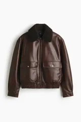 Campera de estilo aviador confeccionada en tejido revestido con acabado de cuero sintético. Presenta cuello de sherpa en contraste, cierre frontal de cremallera, hombros caídos y bolsillos delanteros de parche con solapa y botón a presión. Incluye elástico fruncido en el bajo y puños ajustables con botón a presión.