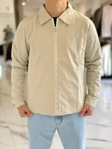 Campera de gabardina beige de corte boxy fit, con cuello camisero, cierre frontal de cremallera blanca y bolsillos laterales ribeteados.