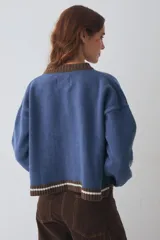 Cardigan de lana color crema con cuello redondo, cierre frontal de botones y mangas largas con puños ajustados. Presenta bordados decorativos en el frente que incluyen un sol, olas y estrellas, con detalles en azul en el cuello y el ruedo.