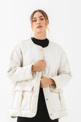 Chaqueta acolchada de color beige con diseño de costuras onduladas, cuello redondo y cierre frontal con botones a presión. Presenta bolsillos frontales con solapa y detalles de tiras ajustables, con un corte holgado y mangas largas.