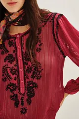 Camisa de manga larga en color negro con tejido semitransparente y detalles de hilos metalizados. Presenta un diseño con bordados florales en tonos tierra, cierre frontal con botones y un lazo decorativo en el cuello.