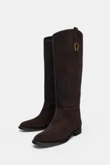 Bota de caña alta confeccionada en serraje, con diseño de estilo hípico. Presenta detalle de pespuntes verticales, tira decorativa con hebilla metálica en la parte superior de la caña y puntera redonda. Cuenta con suela plana de 2,5 cm y sistema de calce tipo pull-on.