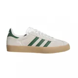 Championes Adidas modelo Gazelle ADV, con diseño clásico de perfil bajo. Presentan una capellada de gamuza en color beige claro, con las icónicas tres tiras laterales y refuerzo en el talón en color verde oscuro. Incluyen cordones blancos, suela de goma color caramelo y el nombre del modelo estampado en dorado en el lateral.