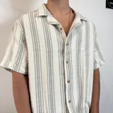 Camisa de manga corta con corte oversize, cuello tipo cubano y cierre frontal con botones. Presenta un diseño de rayas verticales en tonos crudo y gris.