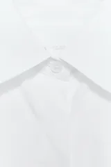 Camisa blanca de popelín de algodón 100%, con cuello solapa, manga larga terminada en puño y cierre frontal con botones ocultos por solapa. Presenta un corte holgado y ruedo curvo.