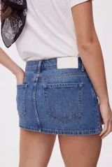 Falda mini de denim azul con calce al cuerpo y tiro medio. Presenta bolsillos delanteros amplios, doble botón en la cintura y aplicaciones de tachas metálicas que recorren la cintura y los bolsillos.