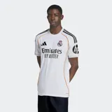 Camiseta de fútbol Adidas del Real Madrid, modelo Primera Equipación temporada 25/26, para hombre. Diseño blanco con detalles en negro (logo Adidas, tres franjas en hombros, texto del sponsor) y ribetes laterales en color naranja/dorado. Incluye escudo del club bordado y logo de sponsor en manga.