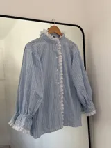 Camisa de rayas verticales celestes y blancas, con cuello alto y cierre frontal de botones. Presenta detalles de puntilla blanca en el cuello, a lo largo de la botonera y en los puños. Tiene mangas largas con terminación acampanada y diseño holgado.