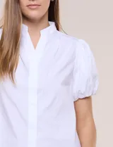 Camisa blanca de calce recto confeccionada en algodón elástico, con cuello mao, abertura frontal en V y cartera con botones ocultos. Presenta mangas cortas abullonadas con puños elastizados y ruedo redondeado.