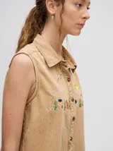 Camisa sin mangas de denim ligero, color celeste lavado, con cuello camisero y botones frontales. Presenta bordados temáticos de playa/mar en el pecho, incluyendo sol, olas, gaviotas, peces y corales/vegetación marina.