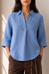 Blusa azul de lino con cuello camisero, escote en V y mangas tres cuartos con vuelta. Presenta un diseño holgado con dobladillo redondeado y un pequeño parche con logo bordado en el lateral inferior.