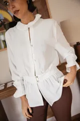 Blusa blanca de manga larga con cuello redondo estilo bebé, cierre frontal con botones y detalle de volados en puños y cintura.