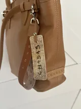 Bolso tipo tote de cuero color marrón claro, con doble asa de mano y correa fina ajustable. Incluye etiqueta colgante con inscripción.