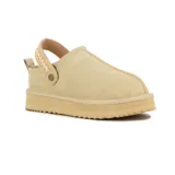 Pantufla tipo zueco color beige, confeccionada en cuero con costura central visible. Presenta una tira trasera ajustable con detalle tejido y suela de plataforma dentada.