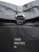 Mochila negra con cierre superior enrollable y hebilla de plástico. Cuenta con un bolsillo frontal con el texto "FORUM INDUSTRIES CO." y un parche en forma de diamante en la parte inferior. Tiene un bolsillo lateral con cierre.
