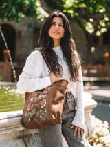 Bolso tipo tote bag confeccionado en cuero serraje color marrón, con diseño artesanal que incluye bordados florales coloridos en el panel frontal. Cuenta con asas de hombro y cierre superior.
