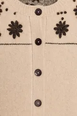 Chaqueta de punto confeccionada en mezcla de lana y alpaca, color beige. Presenta cuello redondo, manga larga, cierre frontal con botones y cinturón ajustable del mismo tejido. Incluye bolsillos delanteros de plastrón y detalles de flores bordadas en color marrón a contraste en la parte superior y en los bolsillos.