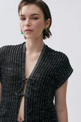 Kimono tejido de punto con hilo de lurex brillante, color negro, con escote en V y cierre frontal mediante lazos. Presenta un diseño sin mangas y textura acanalada.