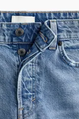 Jean de corte wide leg con diseño de cinco bolsillos, confeccionado en denim de algodón rígido. Presenta un lavado claro con efecto desgastado y un detalle de rotura en la rodilla izquierda. Cuenta con cintura clásica, cierre de botón y bragueta.