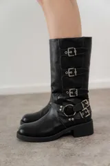 Bota de caña media color negro, con puntera redondeada y taco cuadrado bajo. Presenta tres correas con hebillas metálicas plateadas y un arnés con argolla metálica alrededor del tobillo.