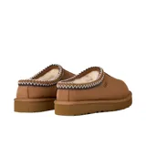 Zuecos UGG modelo Tasman II, confeccionados en gamuza color marrón con forro interno de piel de oveja. Presentan un ribete trenzado decorativo en el cuello y suela de goma resistente.