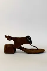 Sandalia de gamuza marrón con taco cuadrado bajo, diseño de tira tipo dedo y aplique decorativo negro brillante en el empeine. Cuenta con tira ajustable en el talón con hebilla.