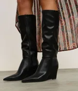 Bota de caña alta Satinato en color negro, confeccionada en material sintético con acabado liso. Presenta un diseño de punta fina y tacón tipo anabela, con una estructura ajustada a la pierna.